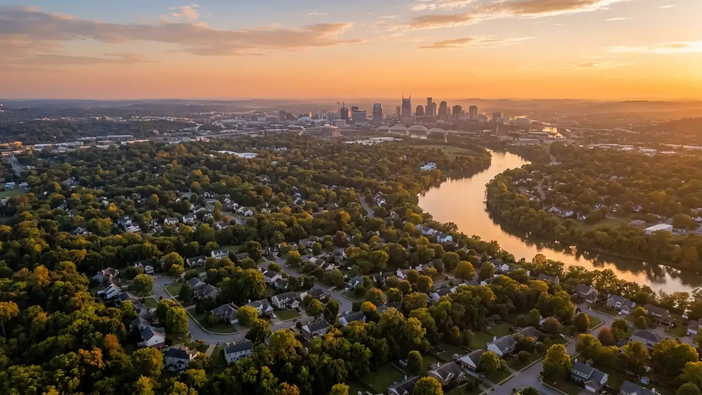A golden sunset over a wide river winding past tree-covered suburban neighborhoods toward a distant downtown skyline
